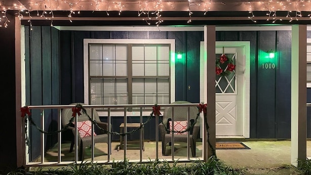 Una casa con porche adornado con luces de cuerda y coronas, el número '1000' en la puerta y un banco con cojín rojo.