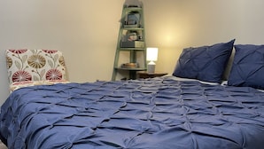 Una cama bien tendida con un edredón azul oscuro, una escalera verde con estantes y una lámpara sobre una mesita de noche.
