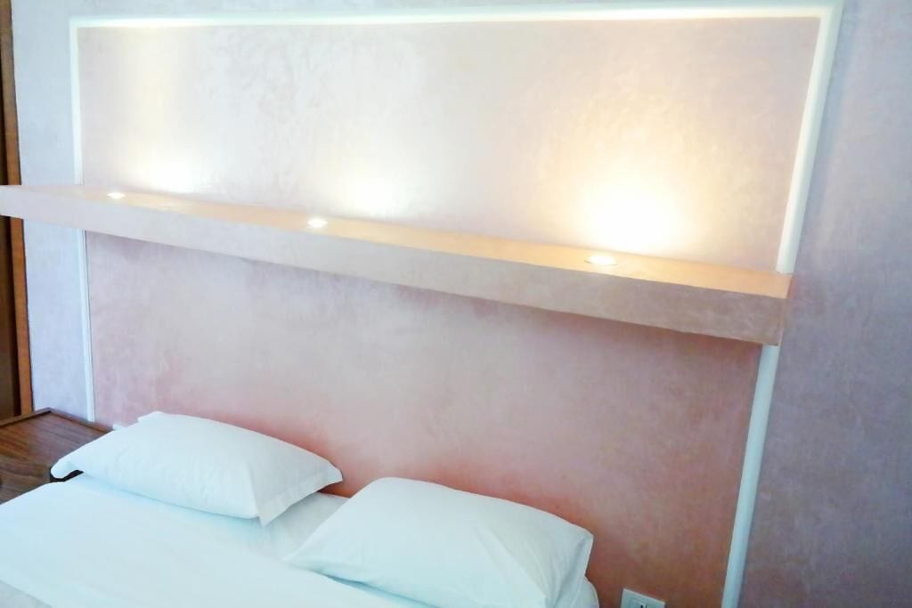 A bedroom with a bed, pillows, a bedside table, and a wall-mounted shelf with lights.
