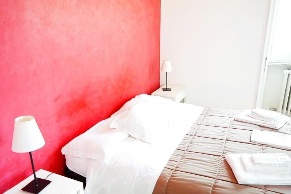 A hotel room with a red accent wall, a bed with white linens, a bedside lamp, and a folded towel on the bed.
