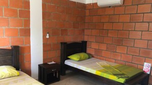 Een kleine kamer met twee bedden, waarvan er een een houten hoofdbord heeft en de ander een eenvoudig hoofdbord. Er is een klein houten nachtkastje met een lade en een papier erop. De muren zijn van rode bakstenen en er is een witte airconditioning unit aan de muur bevestigd.