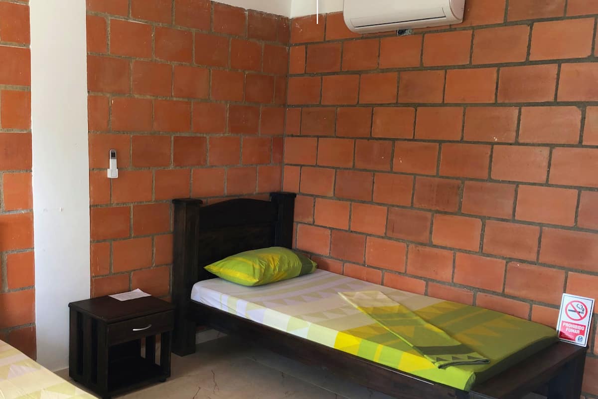 Une petite pièce avec deux lits, l’un doté d’une tête de lit en bois et l’autre d’une tête de lit simple. On y trouve une petite table de chevet en bois avec un tiroir et une feuille de papier posée dessus. Les murs sont en briques rouges, et un appareil de climatisation blanc est fixé au mur.