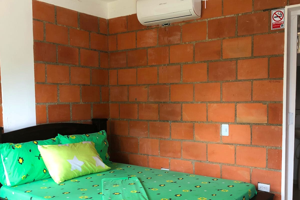 Une pièce avec un mur en briques, un lit recouvert d’une couette à motifs floraux verts, une tête de lit noire et un appareil de climatisation blanc.
