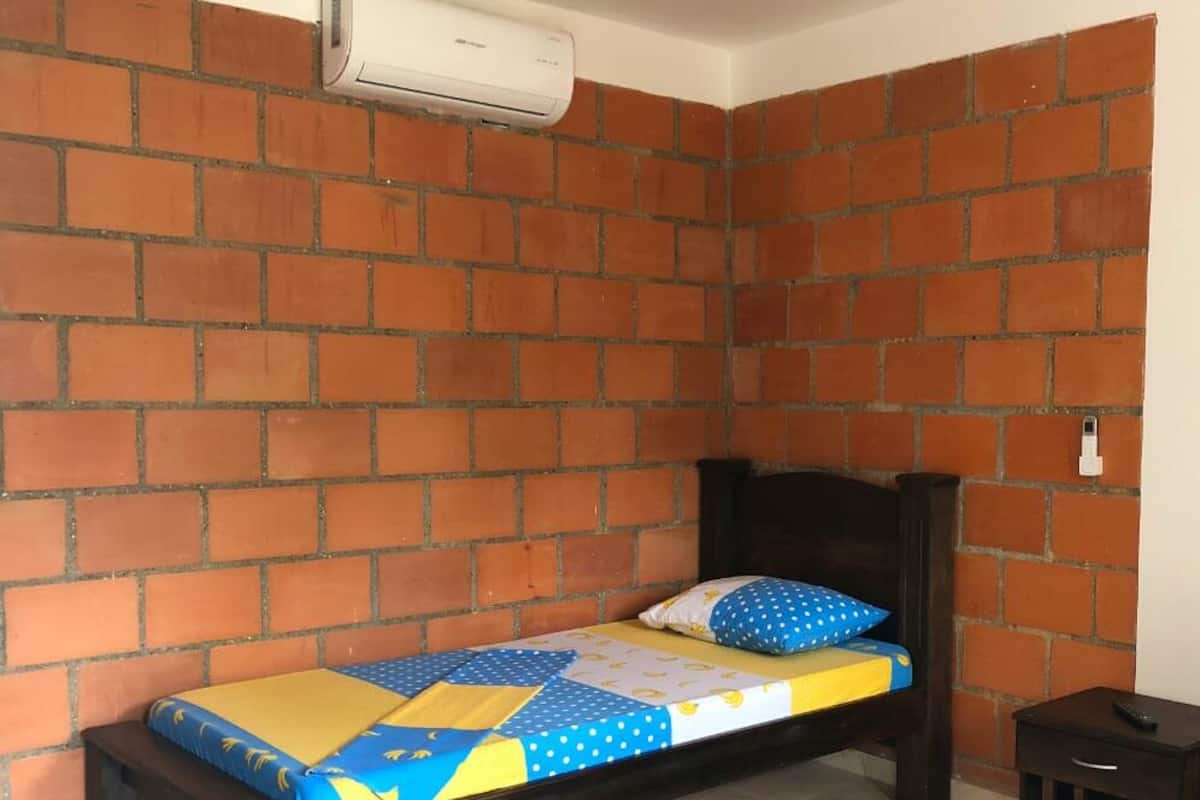 Une petite pièce avec un mur décoré d’un motif en briques, un lit simple recouvert d’une couette bleue et jaune, une table de chevet en bois et une petite table sur laquelle se trouve une télécommande.