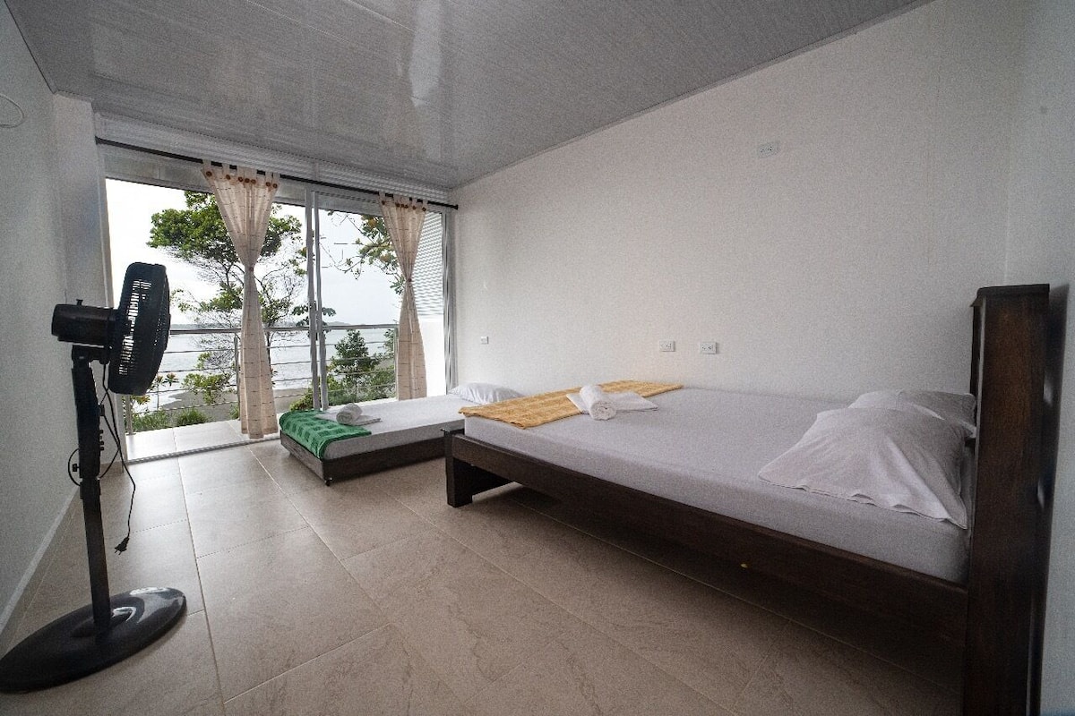 Un dormitorio con cama, un pequeño sofá, un ventilador de pie y una ventana con cortinas.