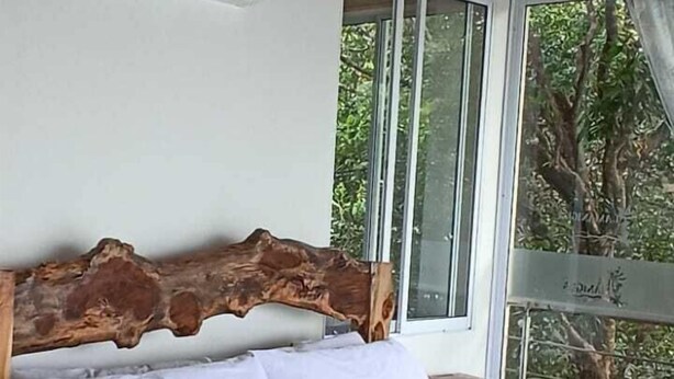 Una cama con cabecera de madera, ropa de cama blanca y ventana con vista a árboles.