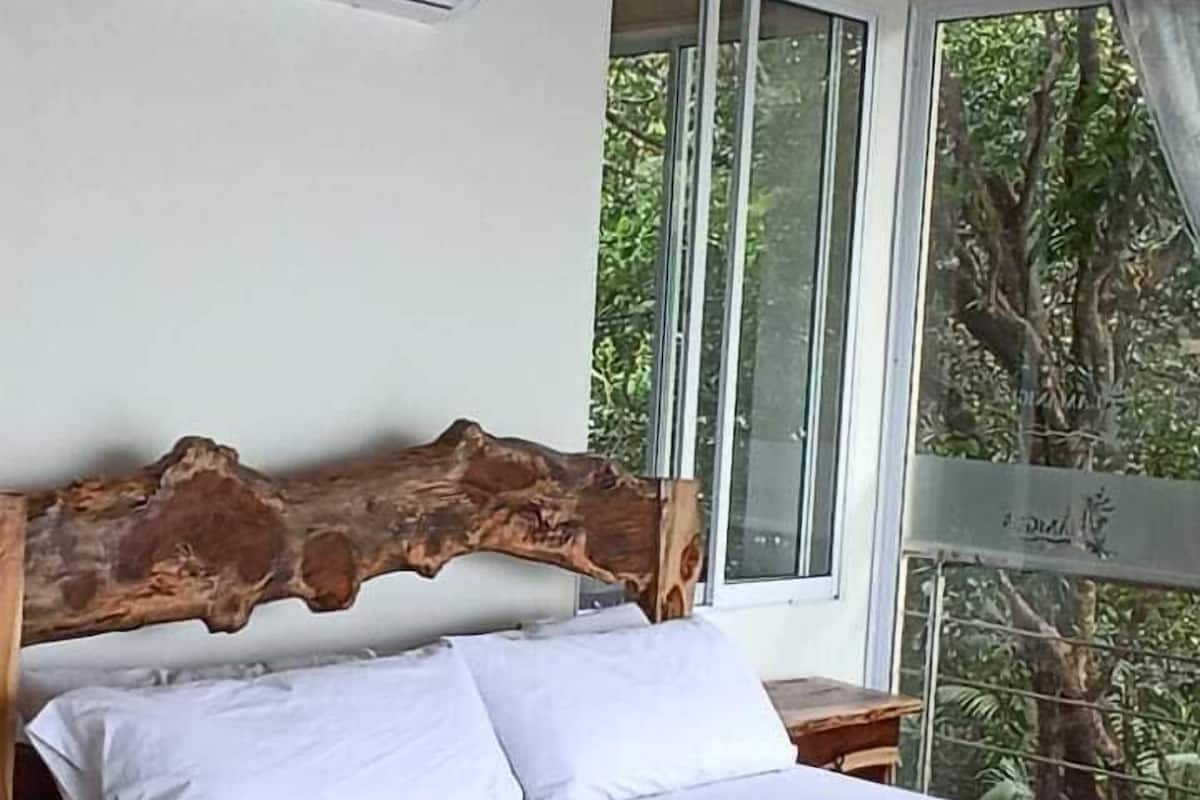 Una cama con cabecera de madera, ropa de cama blanca y ventana con vista a árboles.