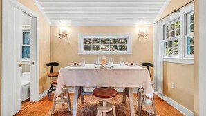 Un comedor con una mesa dispuesta para cuatro, dos sillas a cada lado, un ventanal con vistas a árboles y un techo con vigas blancas.