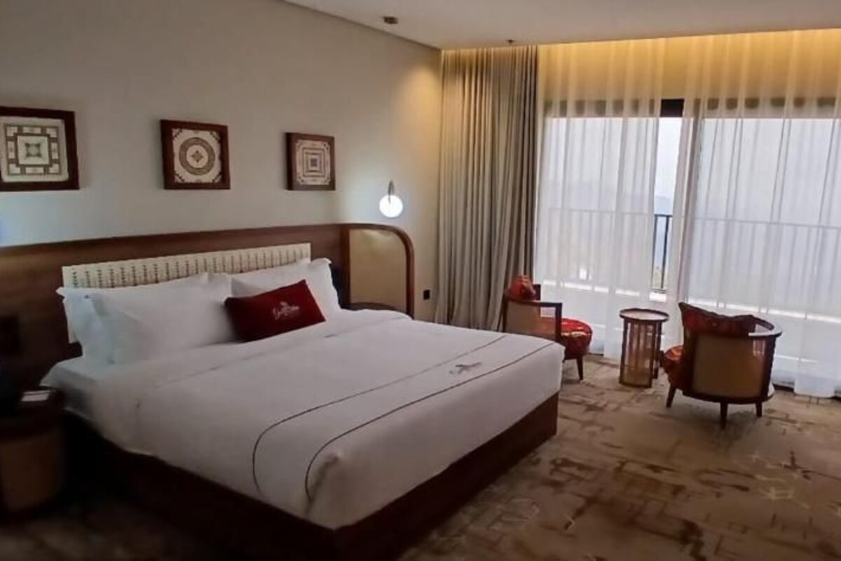 Habitación de hotel con una cama grande, un escritorio, una silla y vistas al exterior a través de amplios ventanales.
