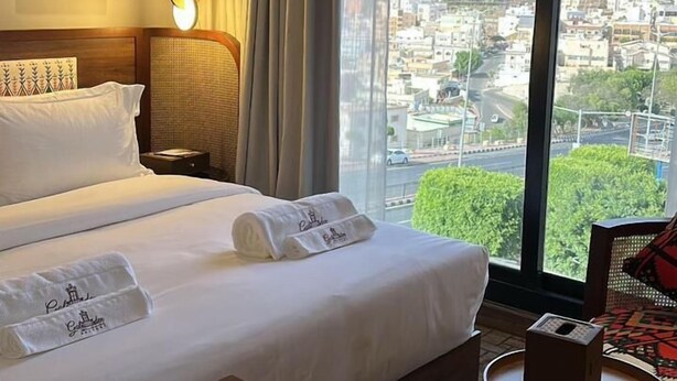 Habitación de hotel con una cama grande, una mesita de noche, una lámpara, una silla y vistas de un paisaje urbano a través de un ventanal amplio.