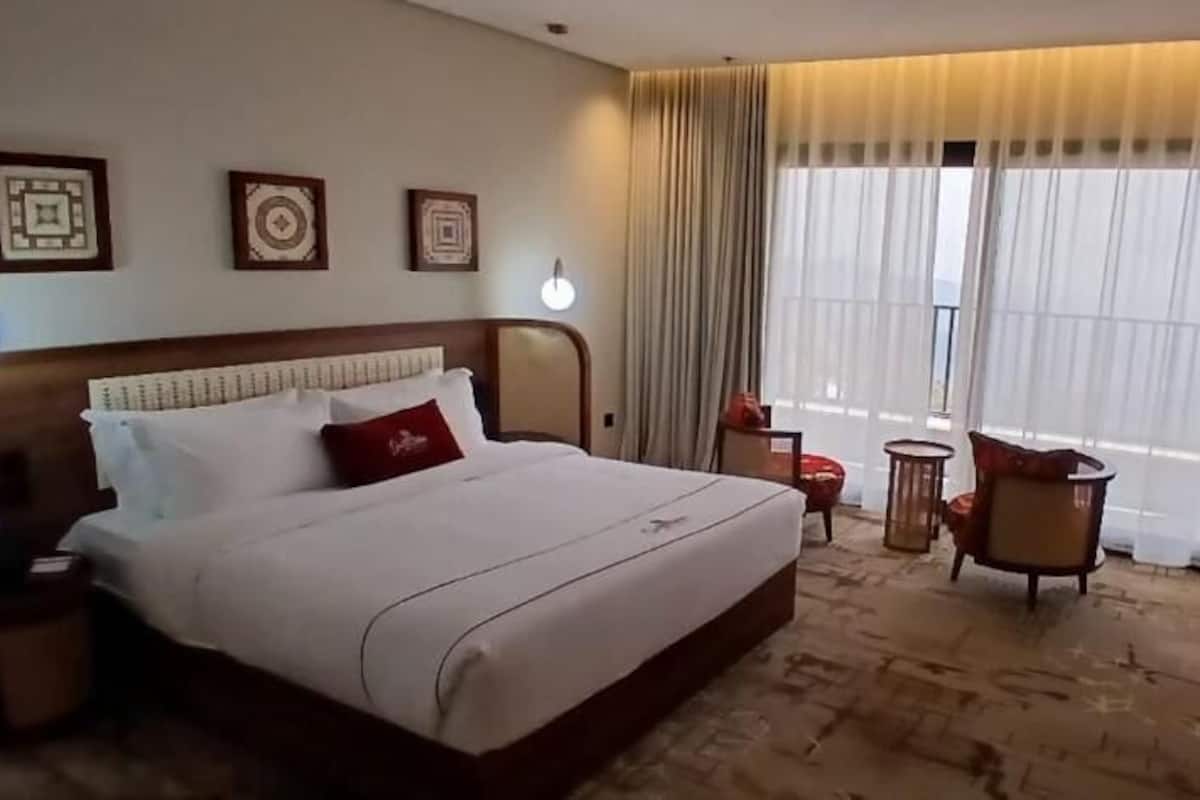 A hotel room with a large bed, a desk, a chair, and a view of the outdoors through large windows.