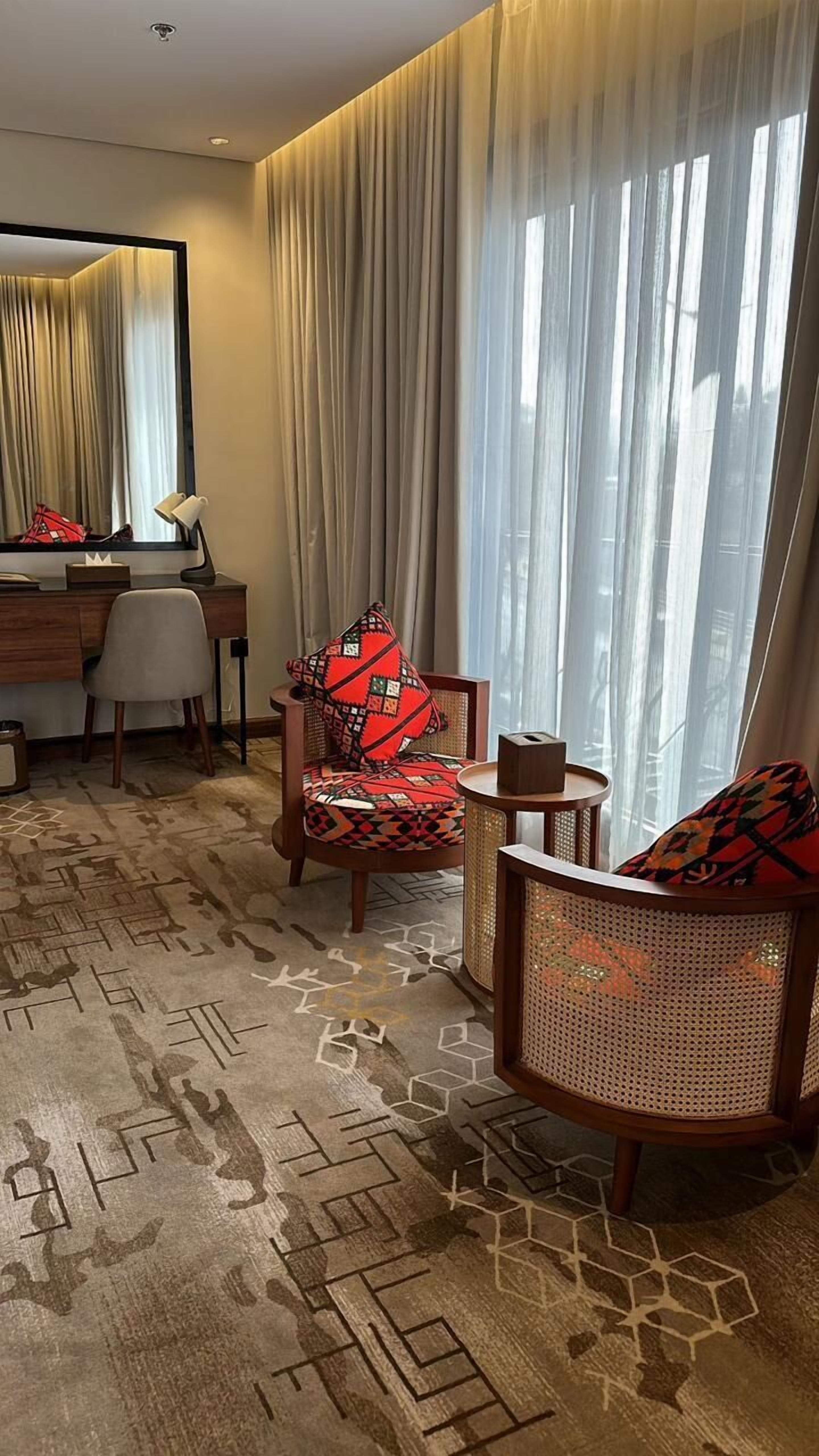 Habitación de hotel con escritorio, dos sillas, una mesita pequeña y un ventanal con cortinas translúcidas.