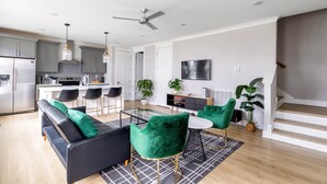 A modern kitchen with stainless steel appliances, a dining area with a glass table and green chairs, a black sofa with green cushions, and a staircase with a white railing.