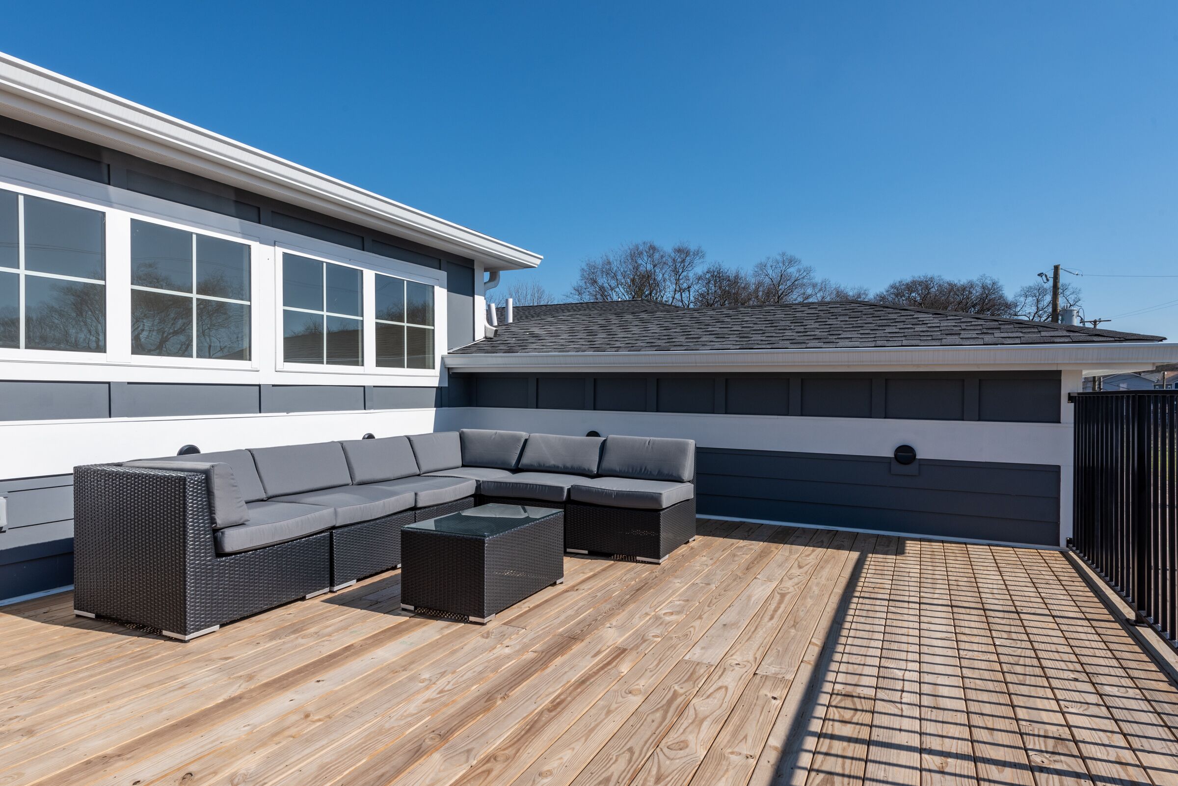 Eine überdachte Terrasse mit Ecksofa und Couchtisch, Holzboden und ein Gebäude mit großen Fenstern und einem dunklen Dach.