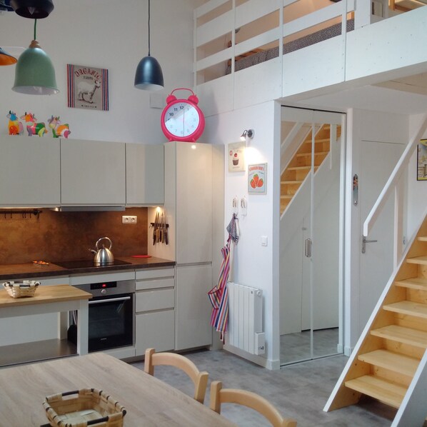 A modern kitchen with a stove, kettle, and hanging utensils. There is a dining table with chairs and a basket. A staircase leads to an upper level.