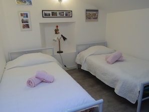 A twin room with two single beds, each with a white duvet and pink towels, a bedside lamp, and a wall-mounted shelf with framed photos.