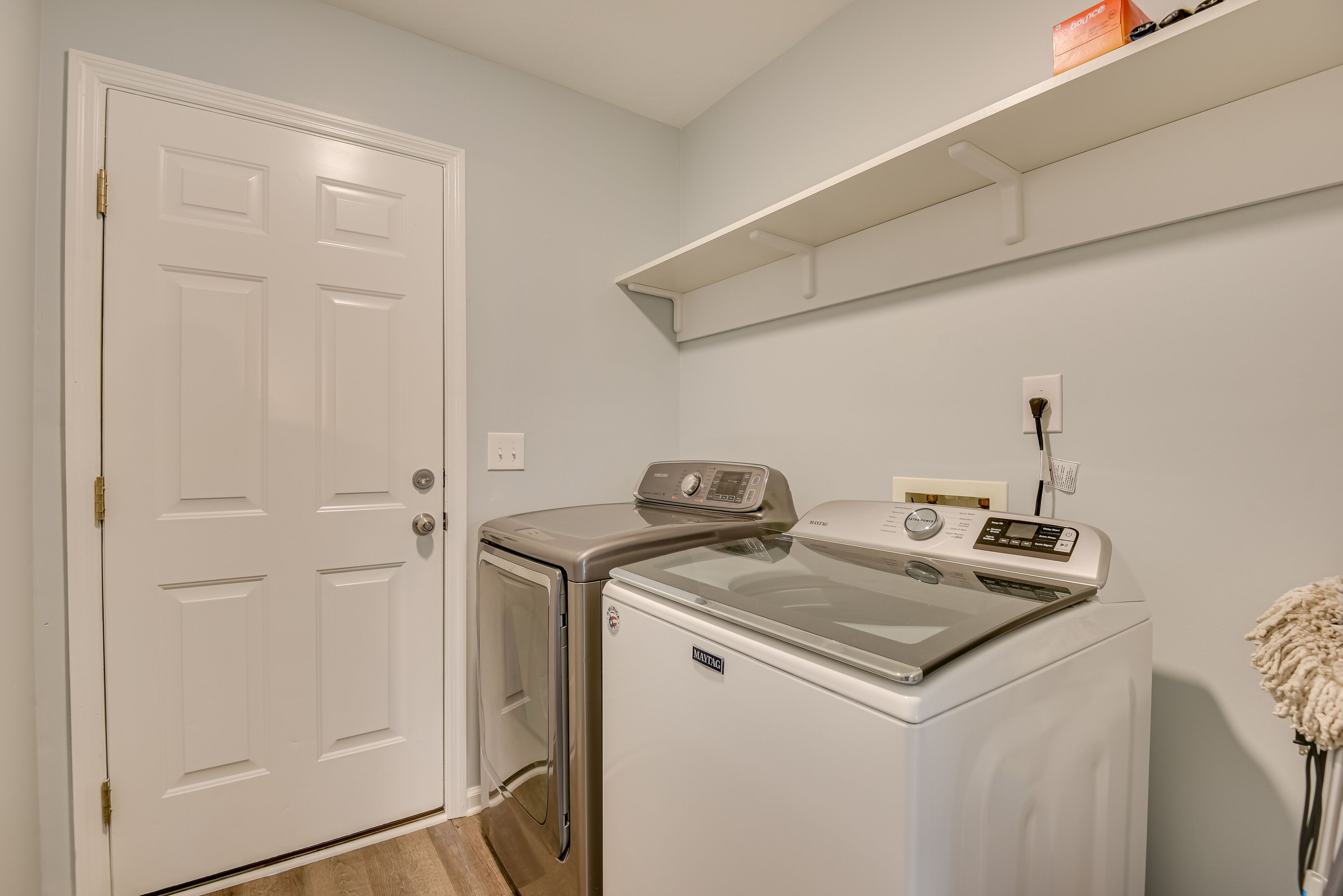 A laundry room with a front-loading washer and dryer, a white door, and a shelf above the dryer.