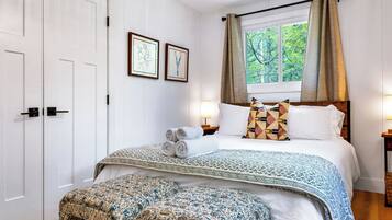 A bedroom with a bed, pillows, a bedside table, a lamp, a window with curtains, and a door.