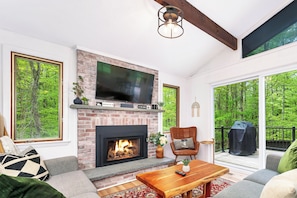 A living room with a fireplace, a television mounted above it, and a large window overlooking a forest.