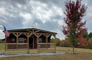 Situated on a grassy area, this cabin features a porch and a tree with vibrant foliage.