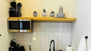 Una cucina compatta con forno a microonde, macchina del caffè e bollitore sul piano di lavoro, un lavello con rubinetto e un armadietto bianco con due ante.