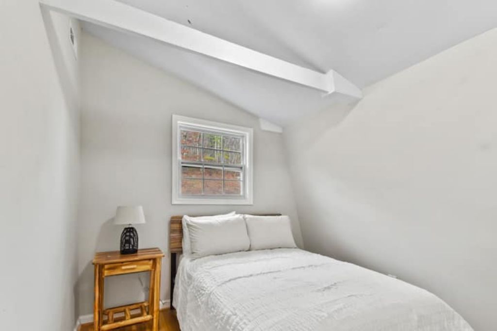 Une chambre avec un plafond en pente, un lit recouvert de linge de lit blanc, une petite table de chevet en bois avec une lampe, et une fenêtre donnant sur un mur de briques.