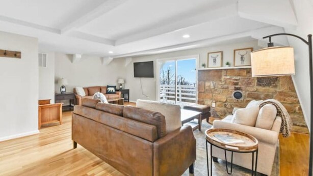 A spacious living room with a wooden floor, a brown leather sofa, a beige sofa, a stone accent wall, and a large window providing a view of the outdoors. - The Overlook (Winchester)