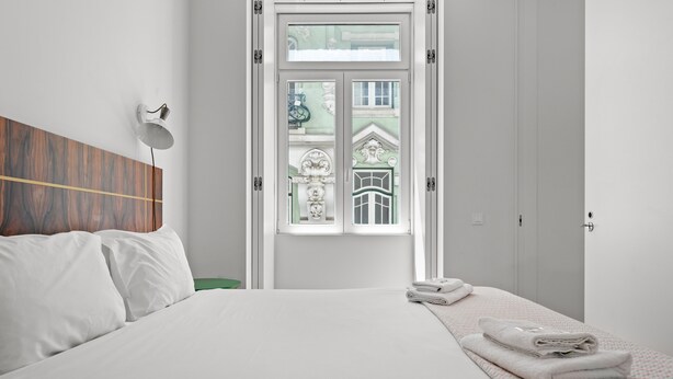 Ett hotellrum med en stor säng, ett grönt sidobord, en vägglampa och ett fönster med utsikt över en byggnad.