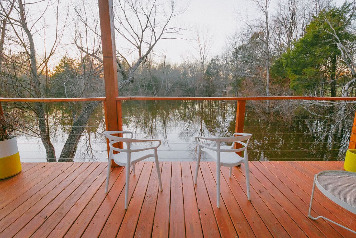 Una terraza de madera con dos sillas blancas orientadas a un lago tranquilo, rodeado de árboles.