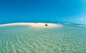 Een strand met helder ondiep water, een zandvlakte en een persoon die op het zand zit.
