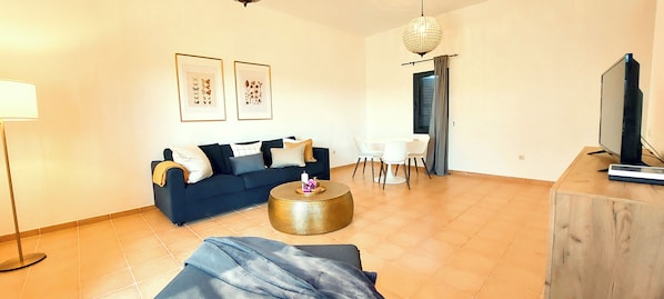 A living room with a dark blue sofa, a round gold coffee table, a TV on a wooden stand, and a dining area with a white table and chairs. - Volcano Sunset View Villa in Corralejo-Tamaragua. (Corralejo City)