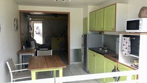 A compact kitchen with green cabinets, a stainless steel refrigerator, and a microwave. There is a wooden table and a chair in the dining area. - Studio La Vigie 3 (Deshaies)
