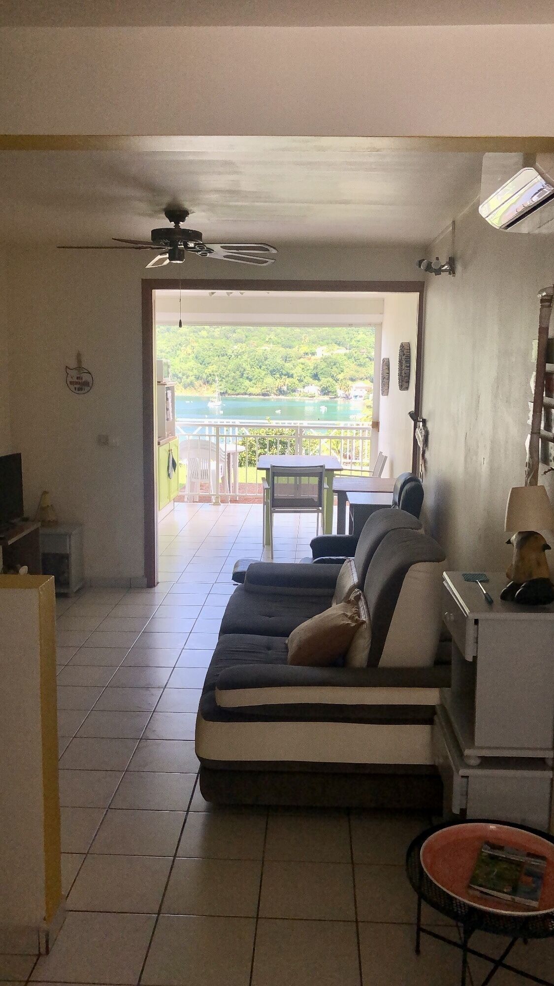 A living room with a sofa, a small table, and a TV. There is a balcony with a view of greenery and a pool.