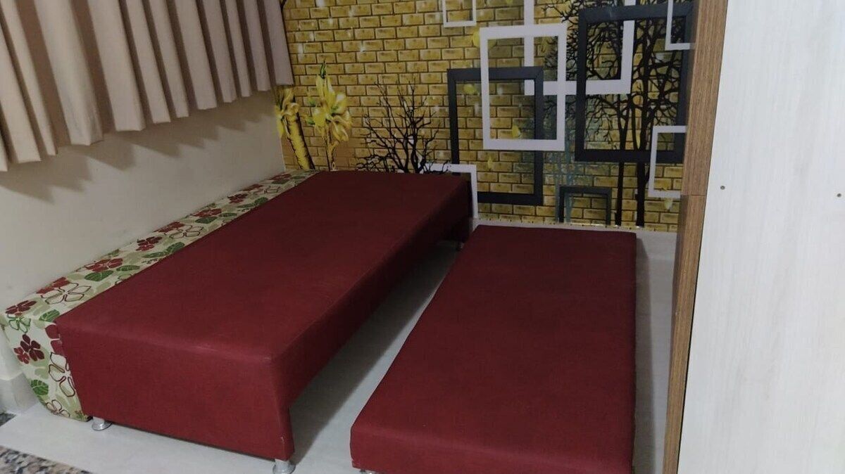 Une pièce avec deux lits recouverts de draps rouges, un mur en briques, une fenêtre ornée de motifs géométriques et une plante décorative.