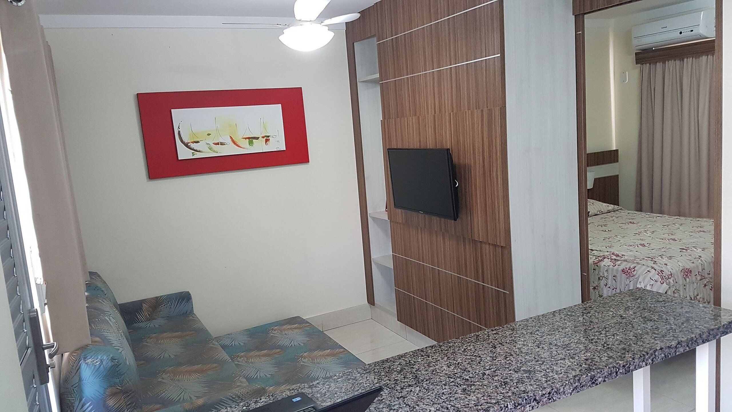 A hotel room with a bed, a TV mounted on the wall, a red-framed artwork, a wooden panel wall, and a small seating area with a bench and cushions.