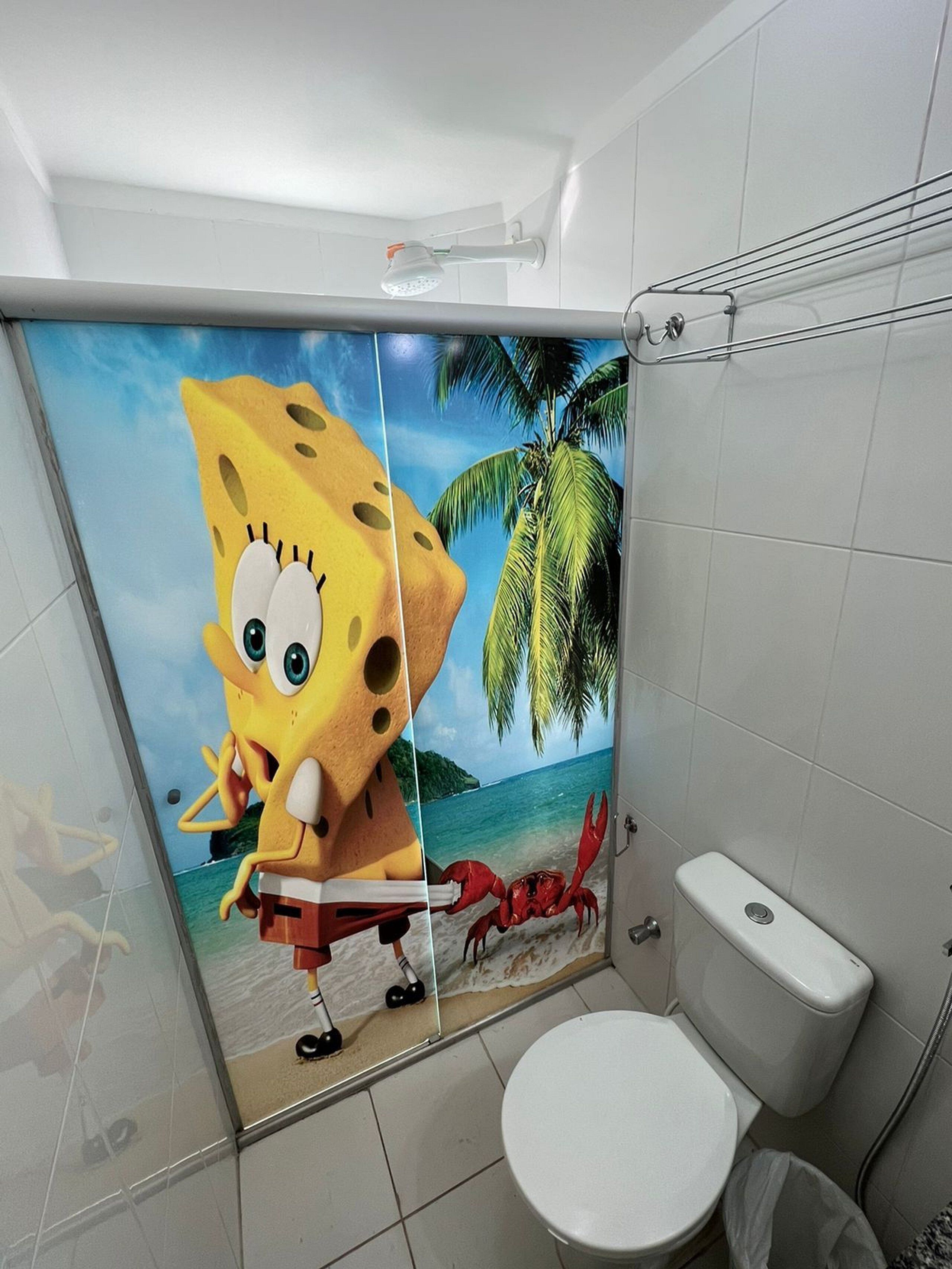 A bathroom with a white toilet, a shower with a glass door, and a wall mural of SpongeBob SquarePants on a beach scene.