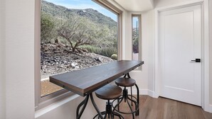 Ein Zimmer mit einem großen Fenster, einem Holztisch, zwei Hocker und einer weißen Tür.