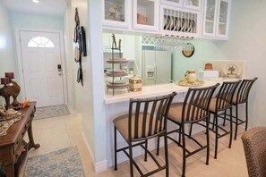 Una cocina con mostrador blanco, un expositor de vidrio de tres niveles y un comedor con cuatro sillas.
