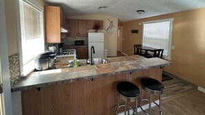 A kitchen with wooden cabinets, a granite countertop, a refrigerator, a microwave, and a sink. There are two bar stools and a dining table with chairs.