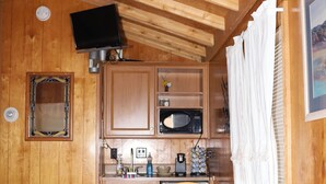 A compact room with wooden walls and ceiling, a bed with a pink blanket, a wooden dining table with chairs, a microwave, a sink, and a television mounted on the ceiling.