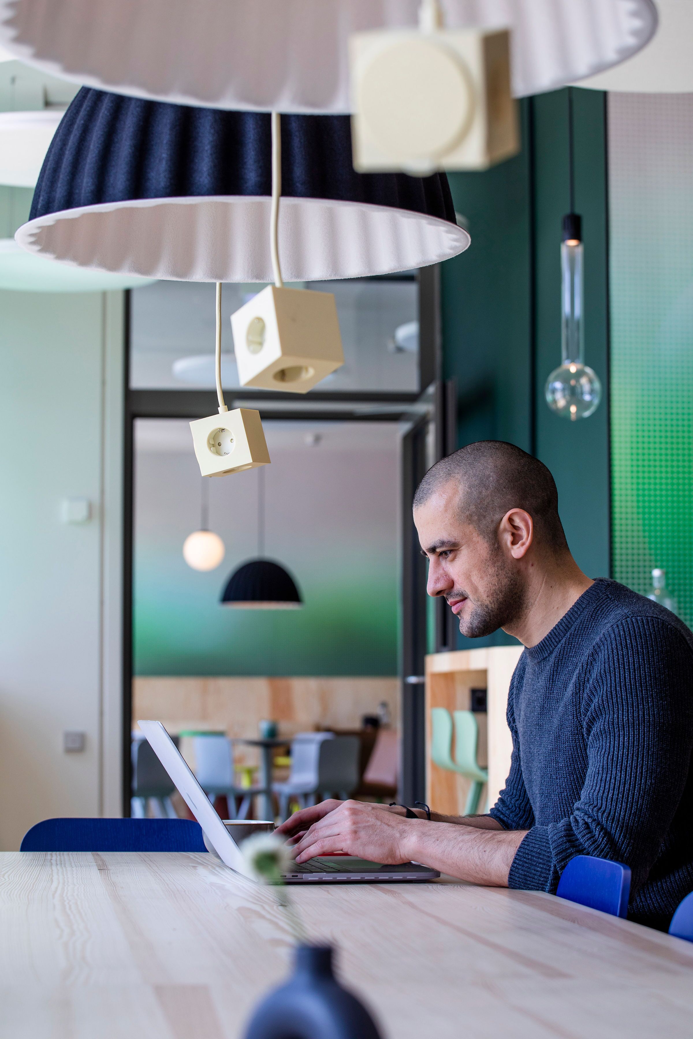 Un hombre trabajando en una mesa con una computadora portátil, en una habitación con iluminación colgante moderna y pared verde.