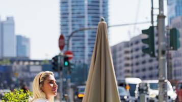 Eine Person sitzt in einem Straßencafé an einem Tisch mit Stühlen, im Hintergrund ein Ampelsignal und ein Gebäude.