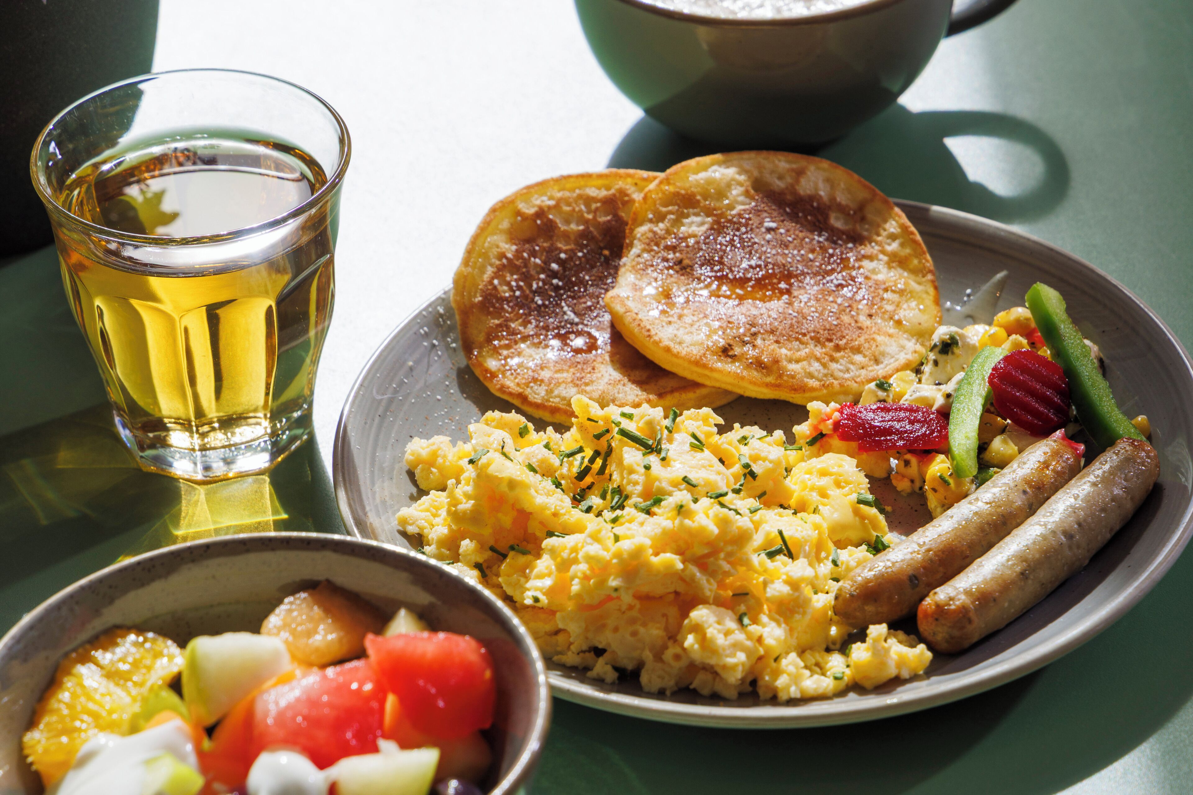Une assiette de petit-déjeuner comprenant des œufs brouillés, des saucisses et des légumes, un verre de thé et un bol de salade de fruits.