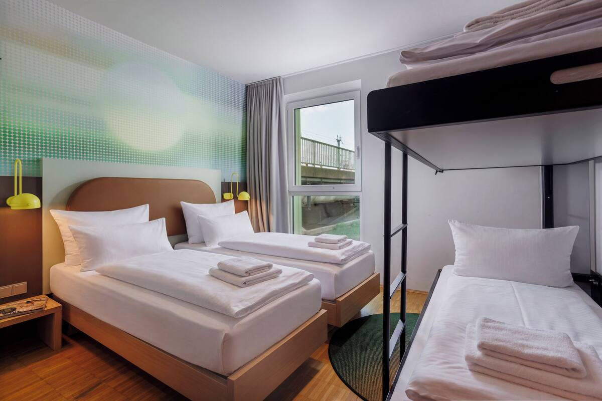 Una habitación de hotel con dos camas, una litera, un ventanal con vistas y una mesita de noche con lámpara.