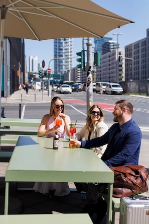 Três pessoas sentadas em uma mesa ao ar livre com bebidas, uma mala e uma bolsa sobre a mesa.