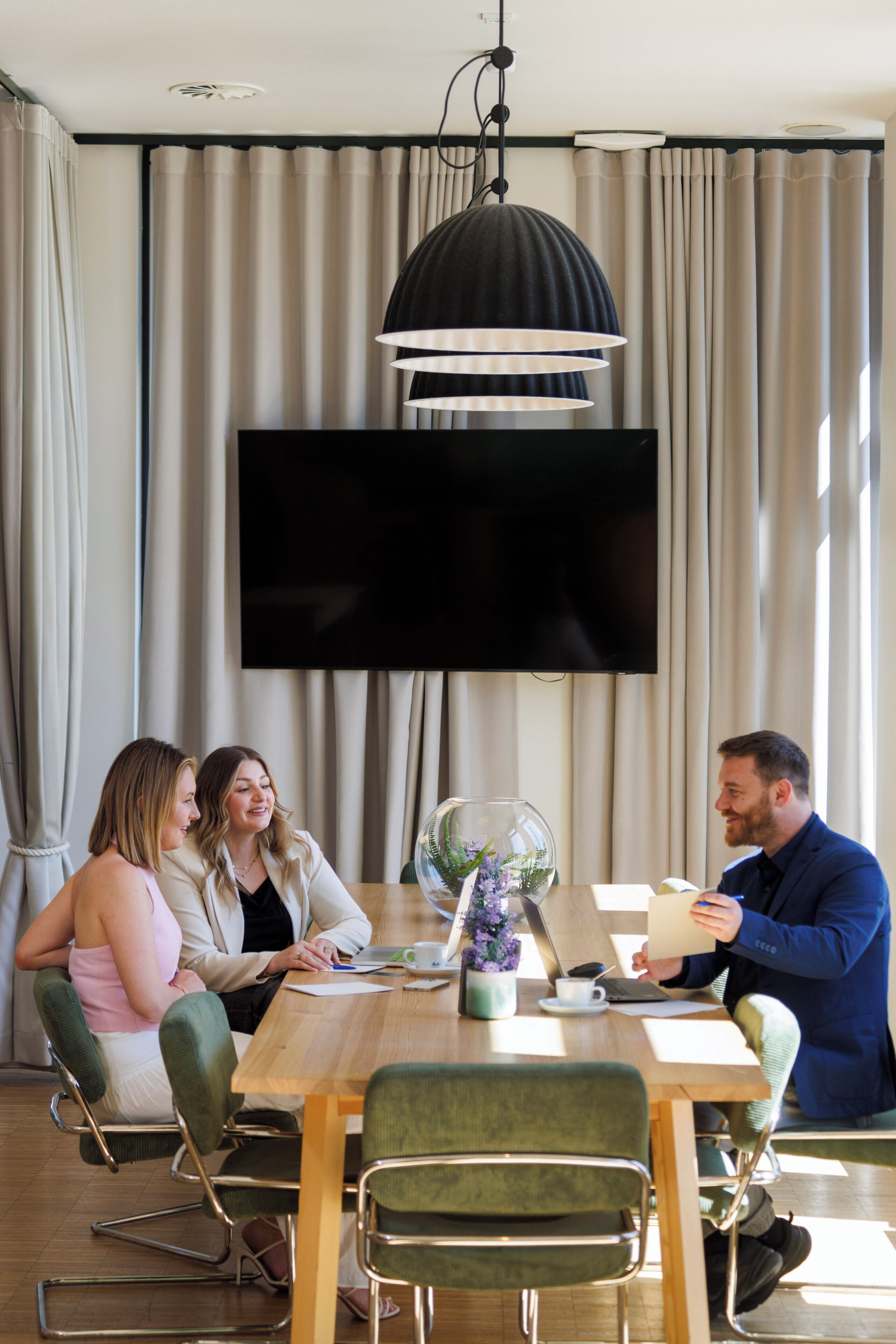 Un espace repas moderne avec une table en bois, des chaises à revêtement vert, un grand téléviseur à écran plat fixé au mur et un luminaire suspendu.