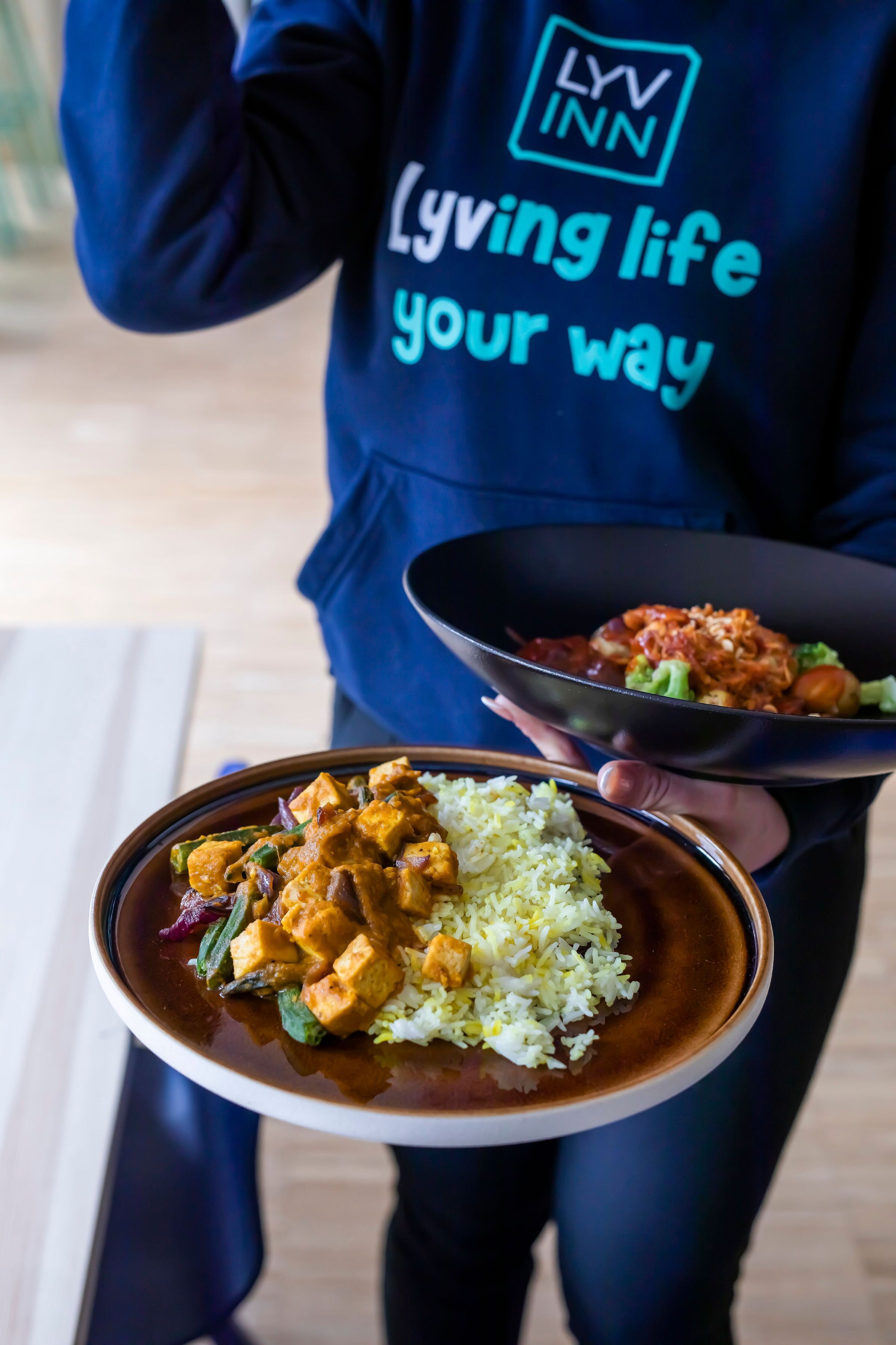A person holding a bowl of curry and rice, wearing a dark blue hoodie with the text "Living life your way".
