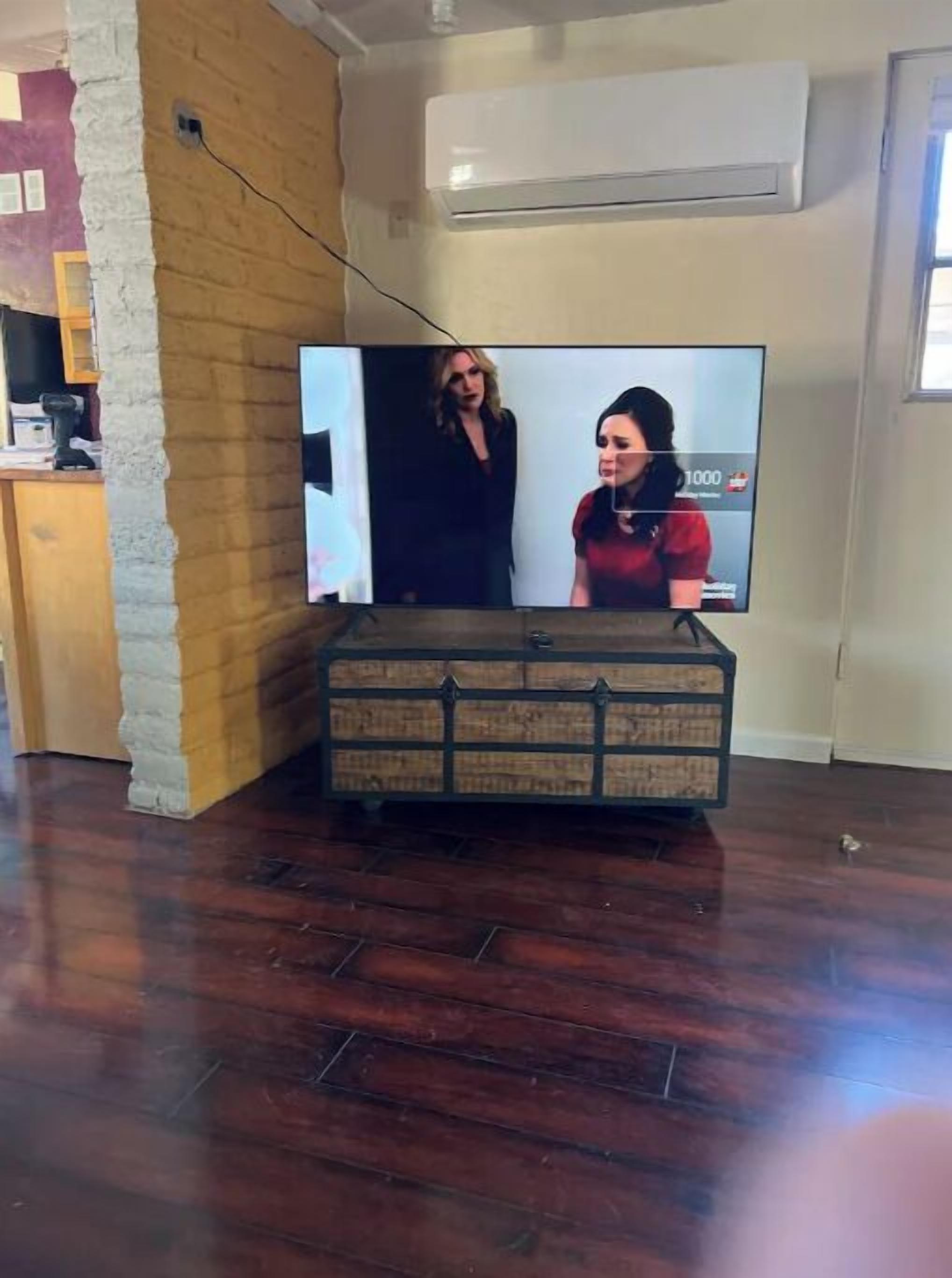 Un televisor sobre un mueble de madera en una habitación con pared de ladrillo y equipo de aire acondicionado.