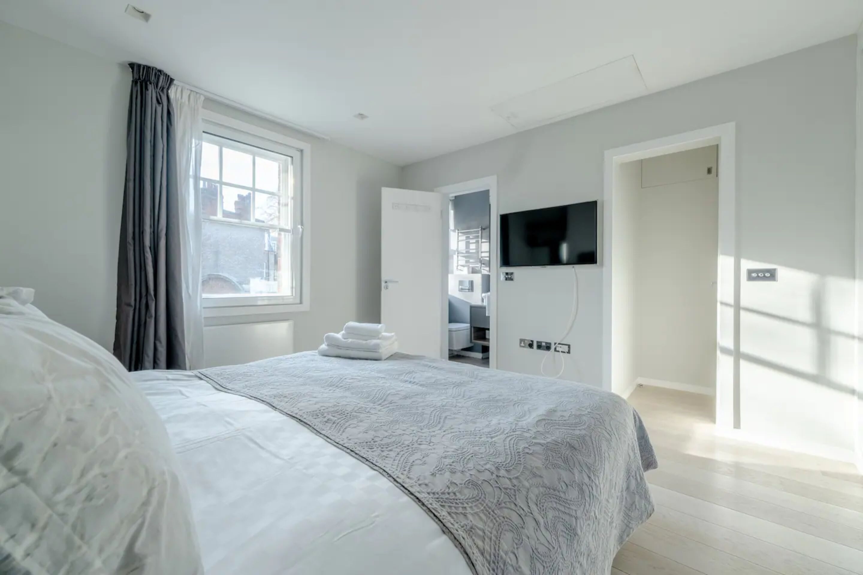 A modern bedroom with a large bed, a TV mounted on the wall, and a view of the outside through a window.