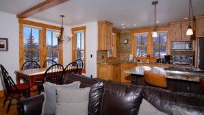 A cozy living room with a leather sofa, wooden dining table, and chairs. A kitchen area with wooden cabinets and a large window is visible in the background. - Perfect spot for your group in Crested Butte! Convenient, walkable, pet friendly (Crested Butte)
